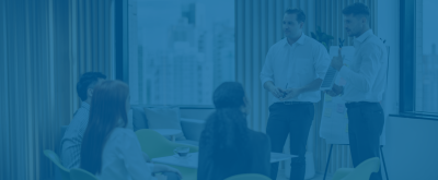 Two men in white shirts lead a presentation for a small group in a modern office with large windows overlooking a city.